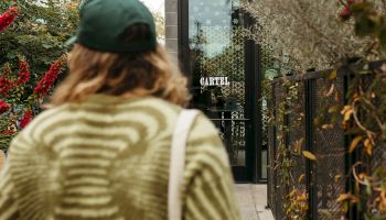 A person wearing a green sweater and cap, carrying a white tote bag, is walking down a pathway towards a building entrance with glass doors.