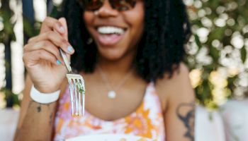 A person with curly hair and sunglasses is smiling and holding a plate of food, ready to eat with a fork in their hand.