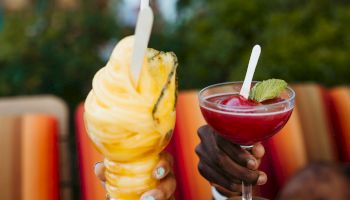 Two hands are holding refreshing drinks: a yellow frozen treat in a cup with a straw and a red cocktail with a lime wedge and a straw.