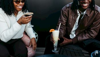 Two people are smiling and enjoying drinks while sitting at a table, with one holding a cocktail and the other a beverage with a straw.