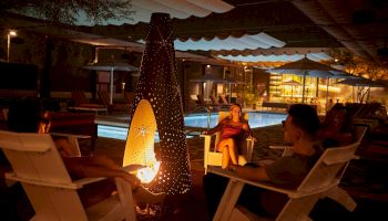 A nighttime poolside lounge with people seated in white chairs around a glowing fire pit, umbrellas, and soft lights in the background.