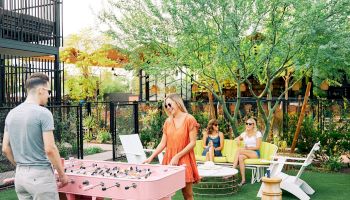 A man watches a pink foosball table outdoors while a woman in an orange dress uses a laptop; two others relax on chairs in a sunny backyard.