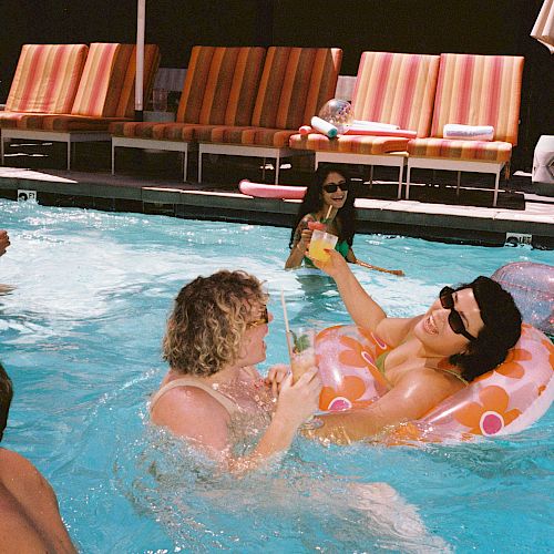 People are swimming and relaxing in a swimming pool, with some sitting on inflatable rings and others by the poolside chairs.