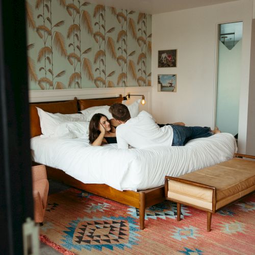 A couple lies together on a bed in a bright, stylish hotel room with patterned wallpaper, a rug, and a doorway to a bathroom, calmly cozy.