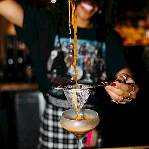 A bartender is pouring a cocktail through a strainer into a coupe glass, smiling in the dimly lit bar.