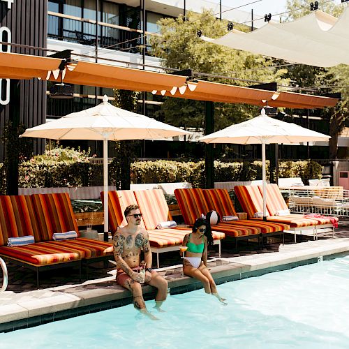 Two people are sitting at the edge of a swimming pool with lounge chairs and umbrellas in the background.