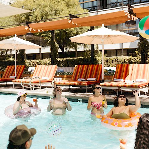 People are in a pool enjoying a sunny day, playing with beach balls and relaxing on floats, surrounded by lounge chairs and umbrellas.