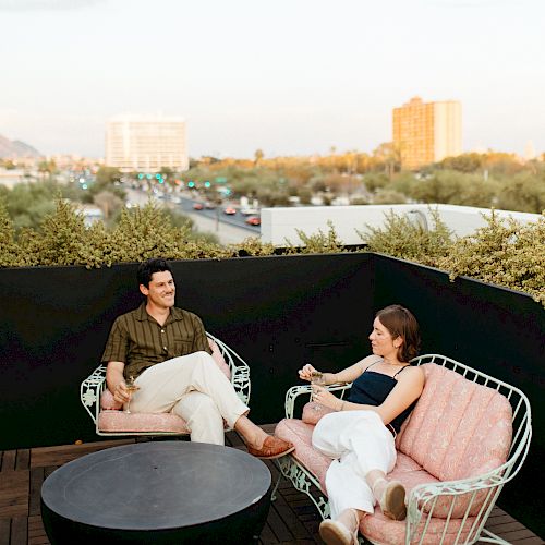 Two people are sitting on a rooftop patio, having a conversation on vintage pink metal chairs, with a cityscape in the background.