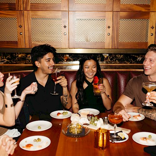 A group of friends is enjoying drinks and food at a restaurant, laughing and chatting around a wooden table.