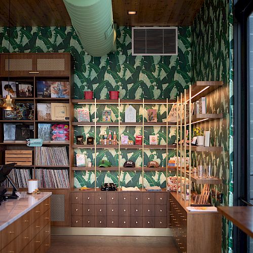 A cozy bookstore or library corner with green foliage wallpaper, shelves full of books, a wooden counter, and drawers underneath the book displays.