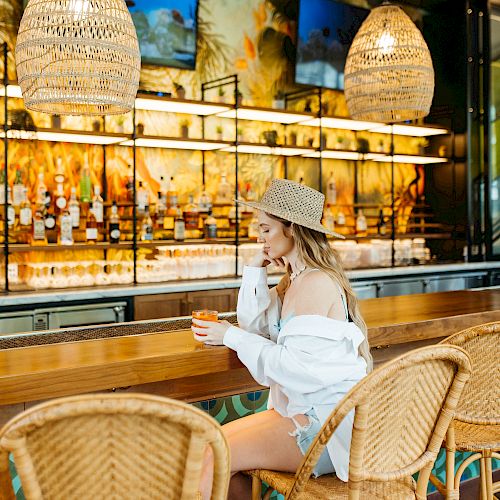 A person wearing a hat and white clothing sits at a bar with hanging lamps and a decorative backdrop, sipping a drink.