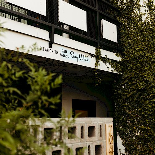 An exterior view of a building with a balcony, greenery, and a concrete block wall. There's a sign with text but the image focus makes it unclear to read.