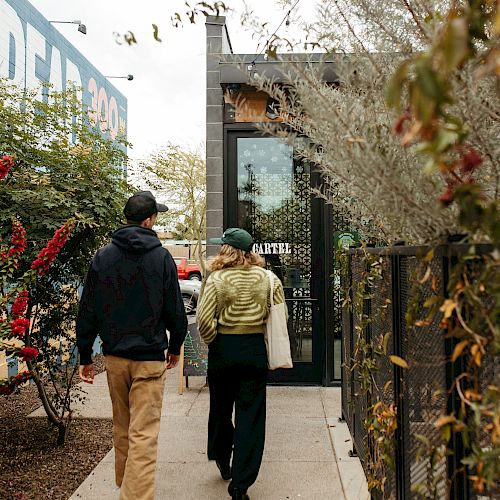 A man and a woman walking down a narrow pathway lined with greenery, approaching a building with a glass entrance.