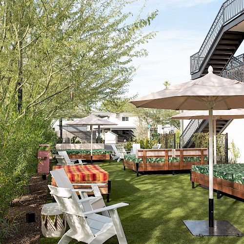 A garden area with Adirondack chairs, an umbrella, plants, and raised beds. There are also stairs and buildings in the background, all on a sunny day.