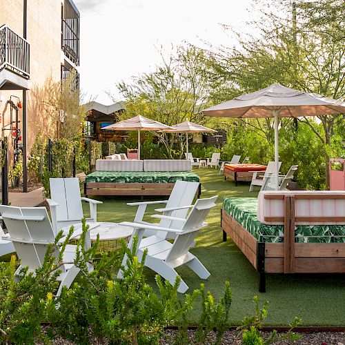 The image depicts an outdoor lounge area with various seating options, including white chairs, daybeds, and umbrellas, surrounded by greenery.