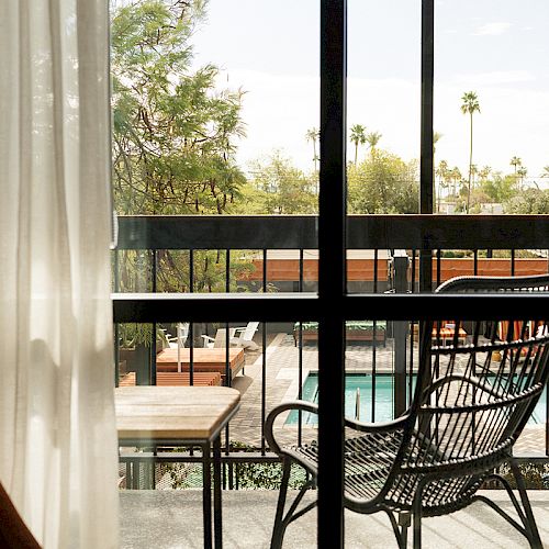 A sunlit balcony with a black chair, small table, and view of a pool area framed by trees, palm trees, and outdoor furniture.