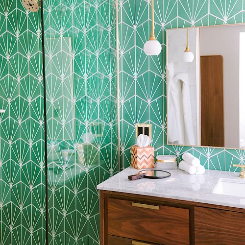 A bathroom featuring green geometric wallpaper, a mirror, pendant lights, a wooden vanity with a marble countertop, and various toiletries.