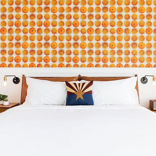 A neatly made bed with white linens, a colorful pillow, wooden headboard, and vibrant orange patterned wallpaper in the background.