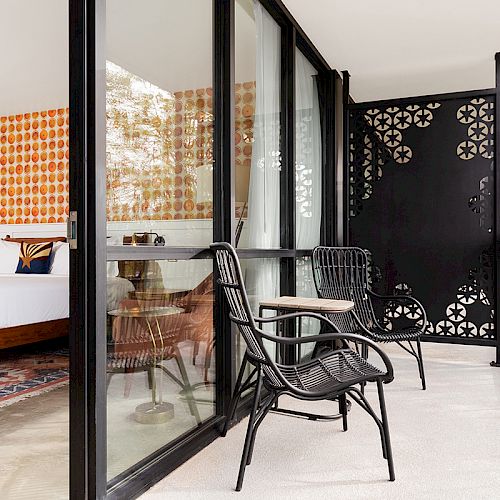 A cozy hotel room with a patterned accent wall and a bed opens to a balcony with two black chairs and a small table, separated by glass doors.