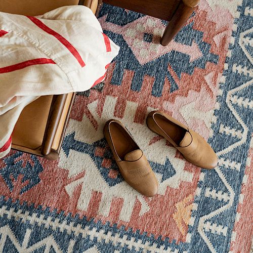 A patterned rug with blue, white, and pink designs, a pair of brown slippers, and part of a chair and white fabric with red stripes are visible.