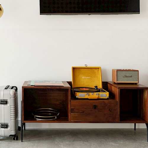 A modern interior setup with a record player, wooden cabinet, small speaker, silver suitcase, and wall-mounted TV, creating a stylish ambiance.