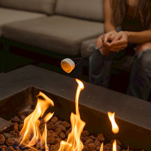 A person roasting a marshmallow over an outdoor fire pit, sitting on a couch.