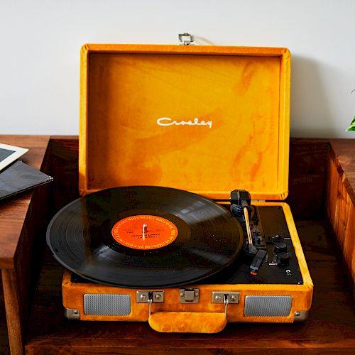 An orange vinyl record player sits on a wooden shelf, with vinyl records beside it and a potted plant nearby.